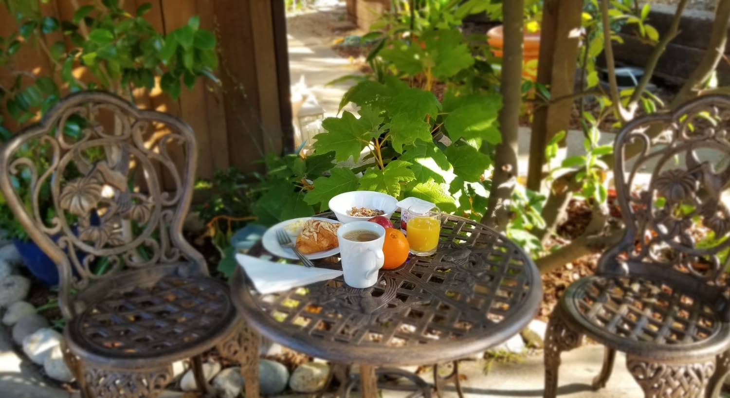 Small breakfast with a pastry, yogurt and granola, orange, apple, apple juice, and coffee in white porcelain dishes sitting on rod iron table on patio