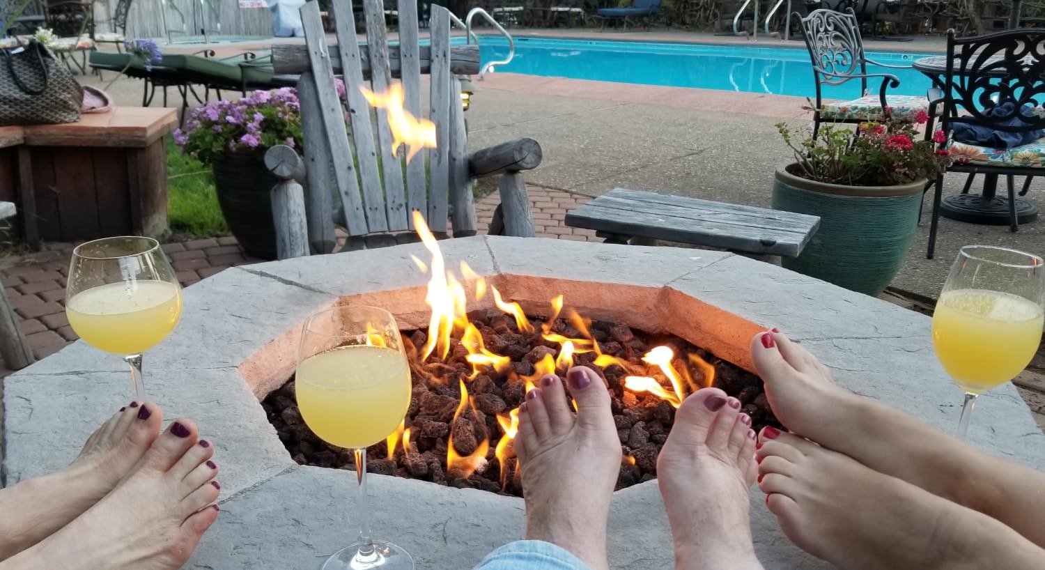 Three ladies feet with painted toe nails resting on firepit with wine glasses overlooking a fire near the pool