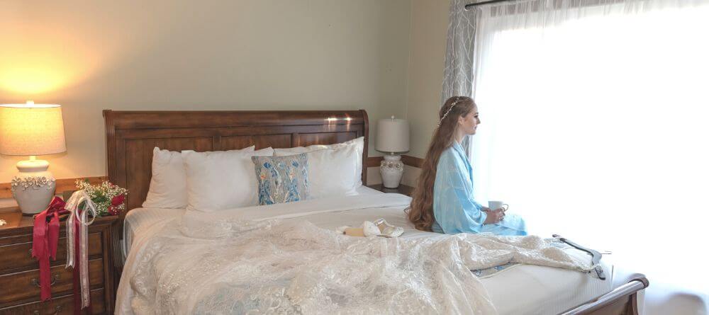 Bride sitting on bed in her robe looking out the window with her wedding dress laid out ready to be put on.