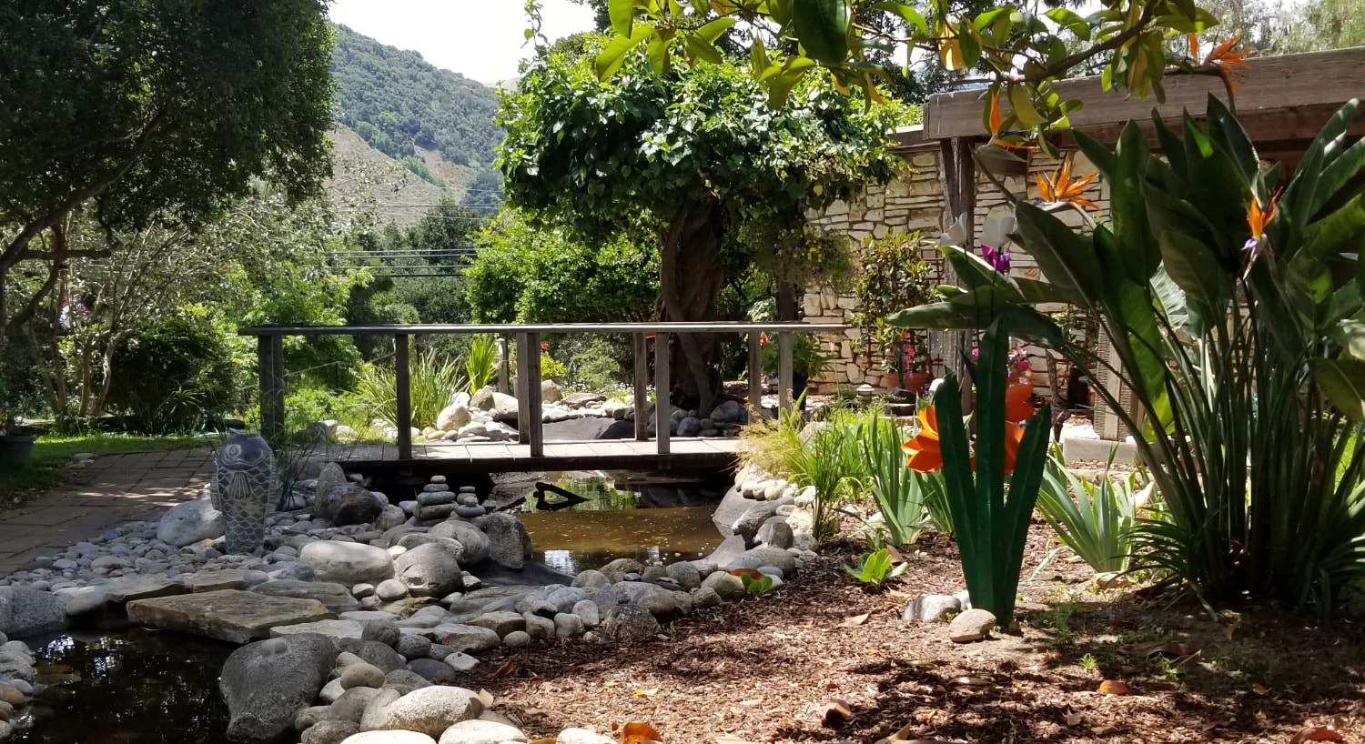 Wooden bridge over small stream surrounded by light gray stones and rocks next to lush garden with plants and orange flowers